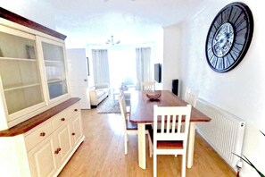 A dining area with a wooden table, white chairs, a white cabinet, and a wall clock. - House Sleeps 5 With Enclosed Garden (Newcastle-upon-Tyne)