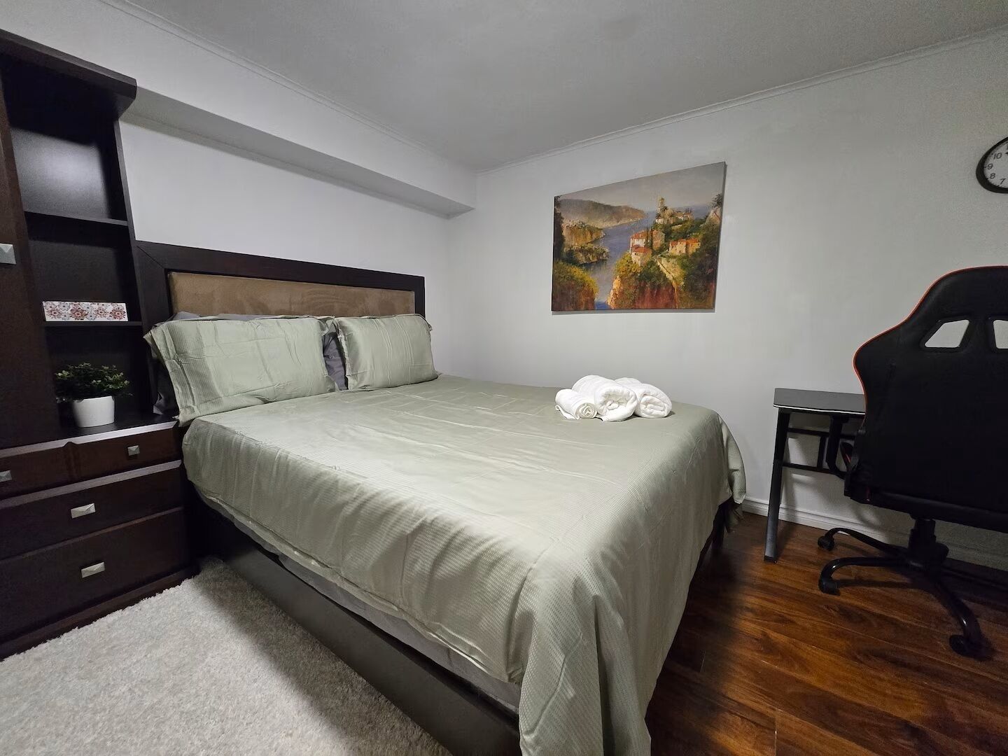 Ein Schlafzimmer mit einem Bett, einem Schreibtisch, einem Sessel, einer Uhr und einem Bild an der Wand.