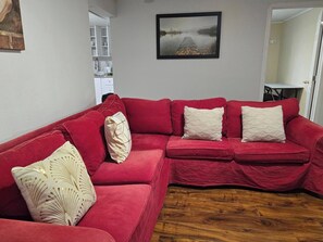 A red sectional sofa with patterned cushions, a framed landscape painting, and a wooden floor. - Two Bedrooms in Basement on Hamilton mountain (Hamilton)