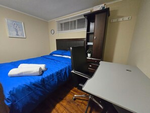 A hotel room with a bed, a desk, a chair, a clock, a window with blinds, and a small potted plant.