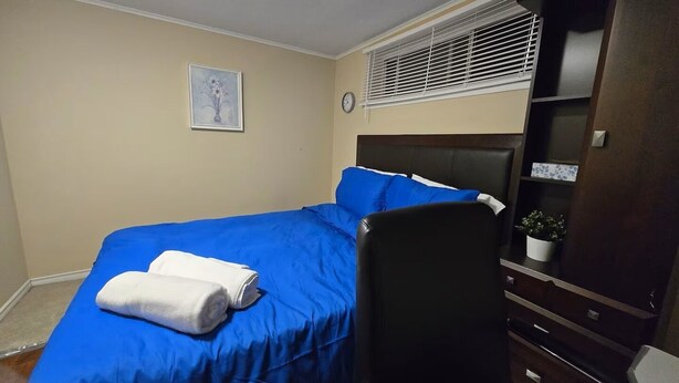 Ein Schlafzimmer mit einem Bett, einem Sessel, einer Kommode, einer Uhr und einem Fenster mit Jalousien.