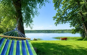 A hammock under a tree by a lake with a boat and a grassy area.
