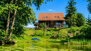 A wooden cabin with a red-tiled roof, situated on a grassy hillside by a calm lake with lily pads.