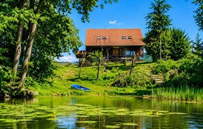 A wooden cabin with a red-tiled roof, situated on a grassy hill by a calm lake with lily pads.