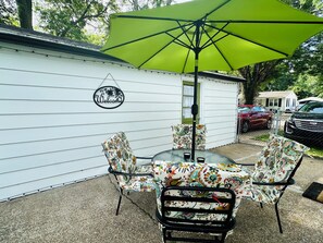 Un patio con una mesa redonda y sillas, una sombrilla verde, y un letrero que dice 'Welcome' en un edificio blanco.