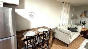 A compact living space with a kitchenette, a dining area with a wooden table and chairs, a sofa, and a lamp. - Cozy Cottage - Pet-Friendly with Fenced Yard (Evansville)