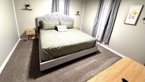 A bedroom with a bed, a bedside table, a small wooden stool, a hanging plant, and a framed picture on the wall. - Cozy Cottage - Pet-Friendly with Fenced Yard (Evansville)