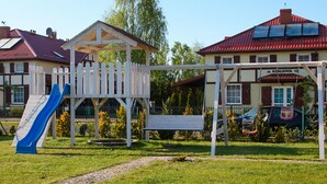 Un parc infantil amb tobogan i cadira d'oscalls, envoltat d'un edifici amb teulada vermella i un rètol que diu 'AZALOWA 13'.