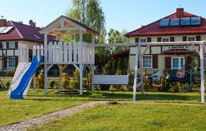 Ein Spielplatz mit Rutsche und Schaukeln, umgeben von einem Gebäude mit rotem Dach und einem Schild mit der Aufschrift 'AZALOWA 13'.