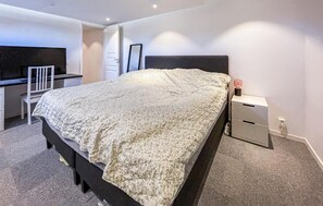 A bedroom with a bed, a desk, a chair, a nightstand, and a wall-mounted TV.