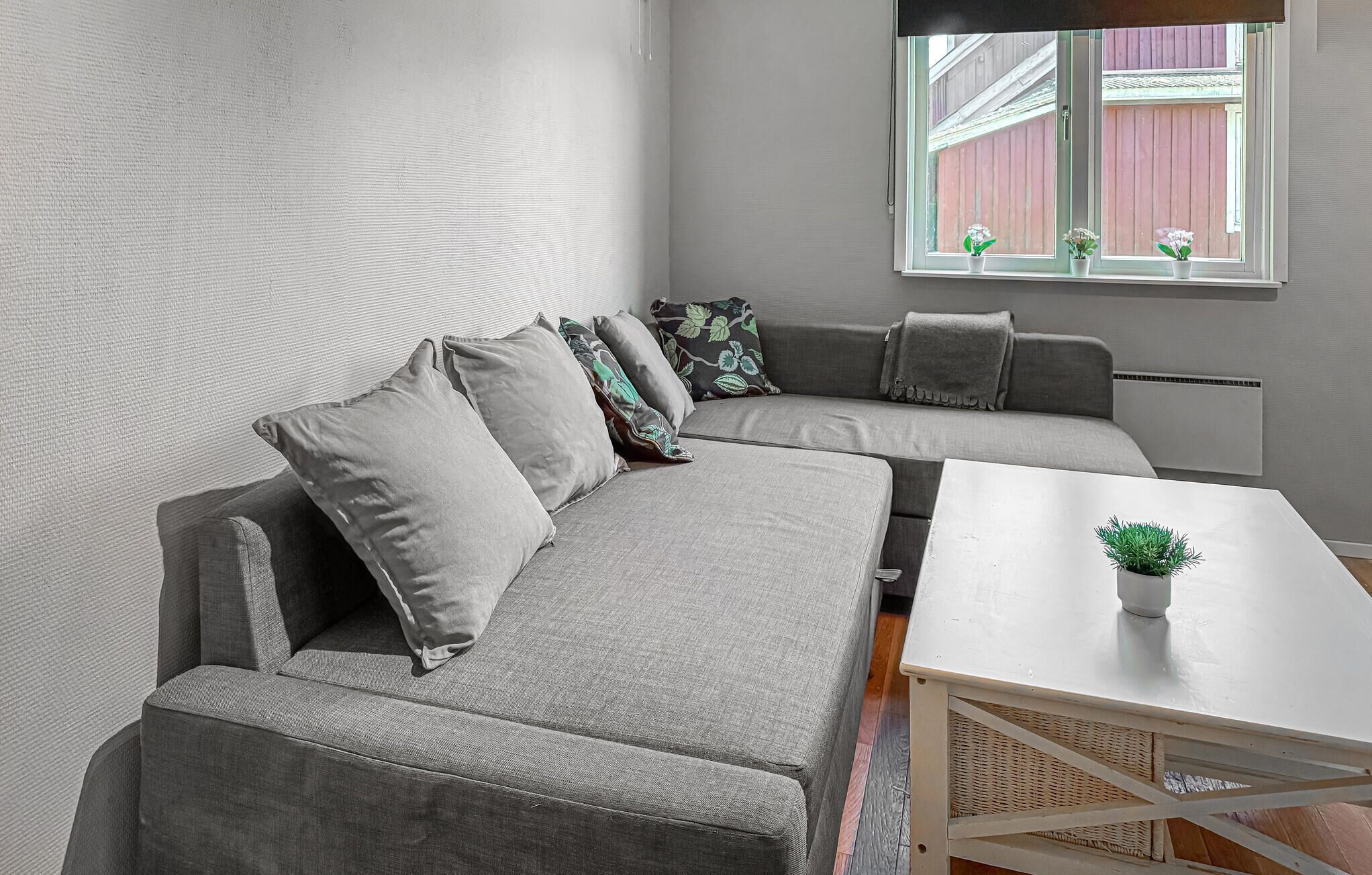 Un salon moderne avec un canapé d’angle gris, une petite table blanche et une fenêtre agrémentée de plantes en pot.