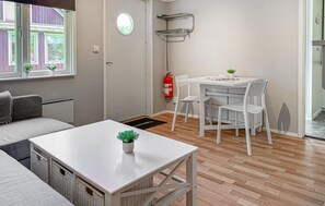 Ein kleiner, moderner Raum mit einem weißen Tisch, zwei Stühlen, einem Sofa, einem Fenster mit Blumentöpfen, einem Feuerlöscher und einer Tür.