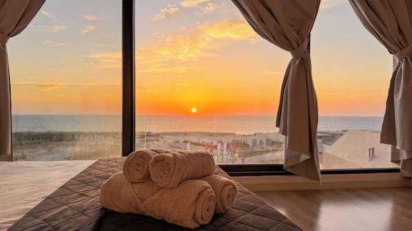 Una habitación con un ventanal grande, una cama con colcha y vistas al atardecer sobre el mar.