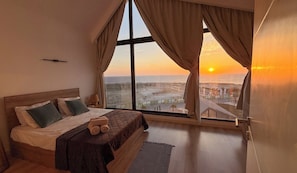 Un dormitorio con una cama grande, dos almohadas, una lámpara de noche y vistas al atardecer sobre un paisaje costero.