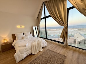 A bedroom with a large bed, a nightstand, a lamp, and a view of the beach and buildings through large windows.
