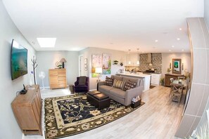 A modern living room with a sofa, ottoman, and a rug. There is a wooden cabinet, a purple armchair, and a dining area with a table and chairs.