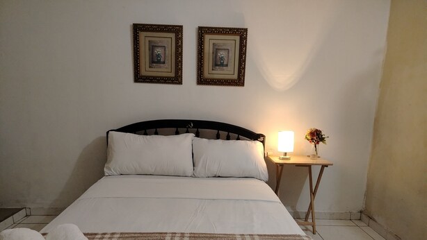 A neatly made bed with white linens, a brown blanket, and two rolled towels. There is a bedside table with a lamp and a vase of flowers. Two framed pictures are hung on the wall above the bed. - Mary's little house (San Juan Bautista)