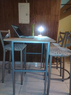 A dining area with a table, two chairs, a laptop, a vase with flowers, and a wall-mounted light fixture.