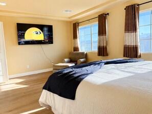 Ein Schlafzimmer mit einem Bett, einem an der Wand montierten Fernseher, einem Fenster mit Vorhängen und einem Sessel.