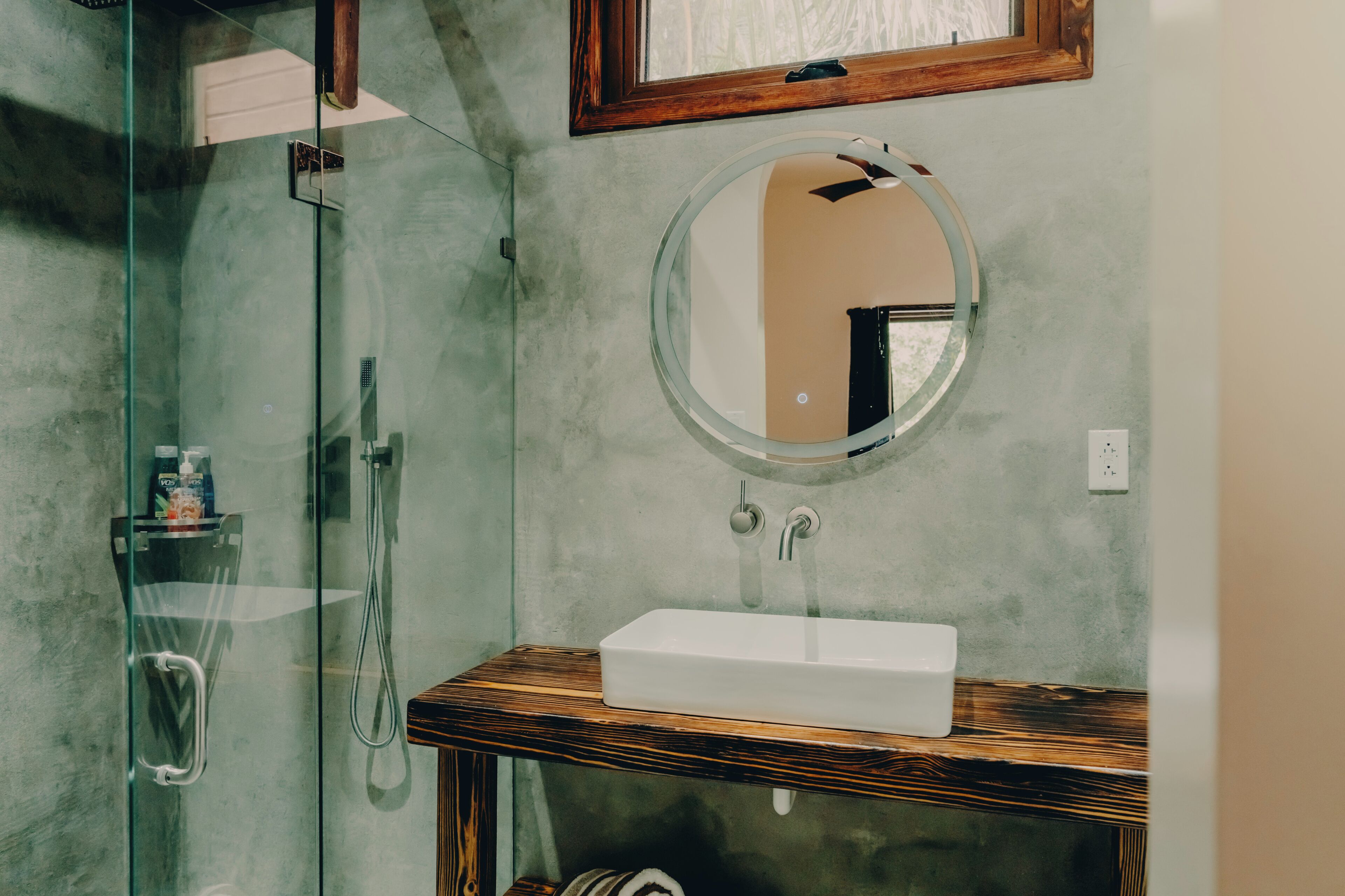 Un baño con cabina de ducha de vidrio, tocador de madera con lavamanos blanco y espejo redondo.