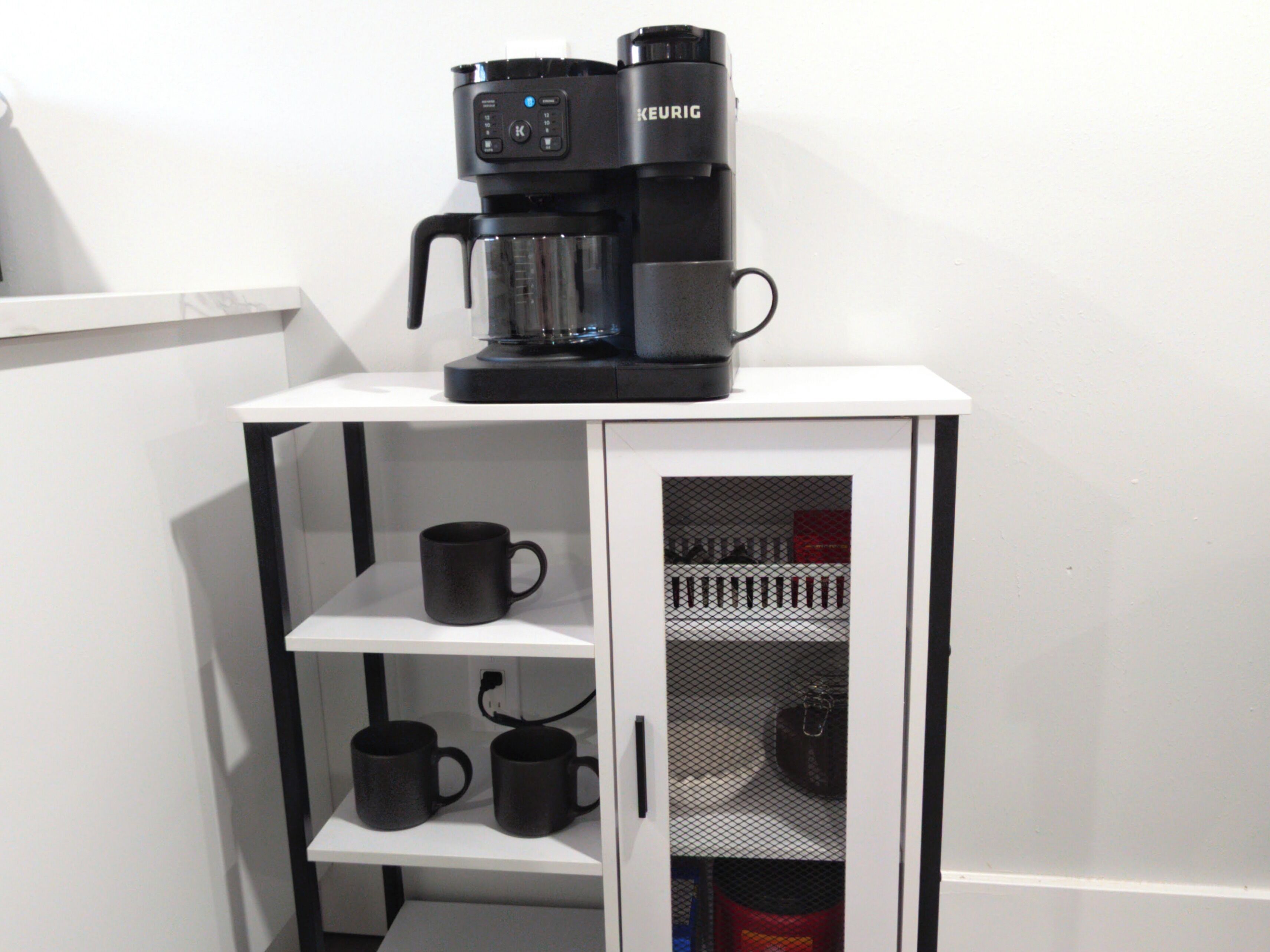 Een kitchenette met een koffiezetapparaat, twee zwarte mokken op een plank, en een gaasdeur.