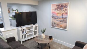Un salon avec un téléviseur à écran plat posé sur un meuble blanc, une table basse ronde ornée d’une plante et un tableau représentant un arbre accroché au mur.