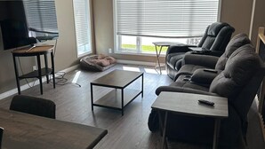 A living room with a sofa, a TV, a coffee table, and a dining area. - Sleep & Grill | 5BR, Huge Yard, Smoker, BBQ (Fox Creek)