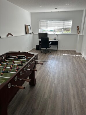 A room with a foosball table, a desk with a computer, and a window with blinds.
