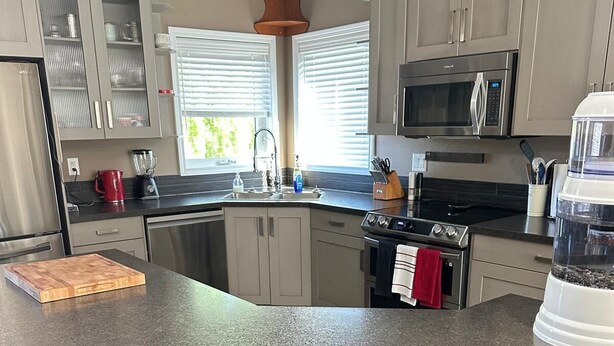 A kitchen with a granite countertop, stainless steel appliances, and a wooden chair. - Sleep & Grill | 5BR, Huge Yard, Smoker, BBQ (Fox Creek)