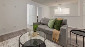 Una sala de estar con un sofá gris, una mesa de centro de vidrio, una planta y una ventana con persianas.