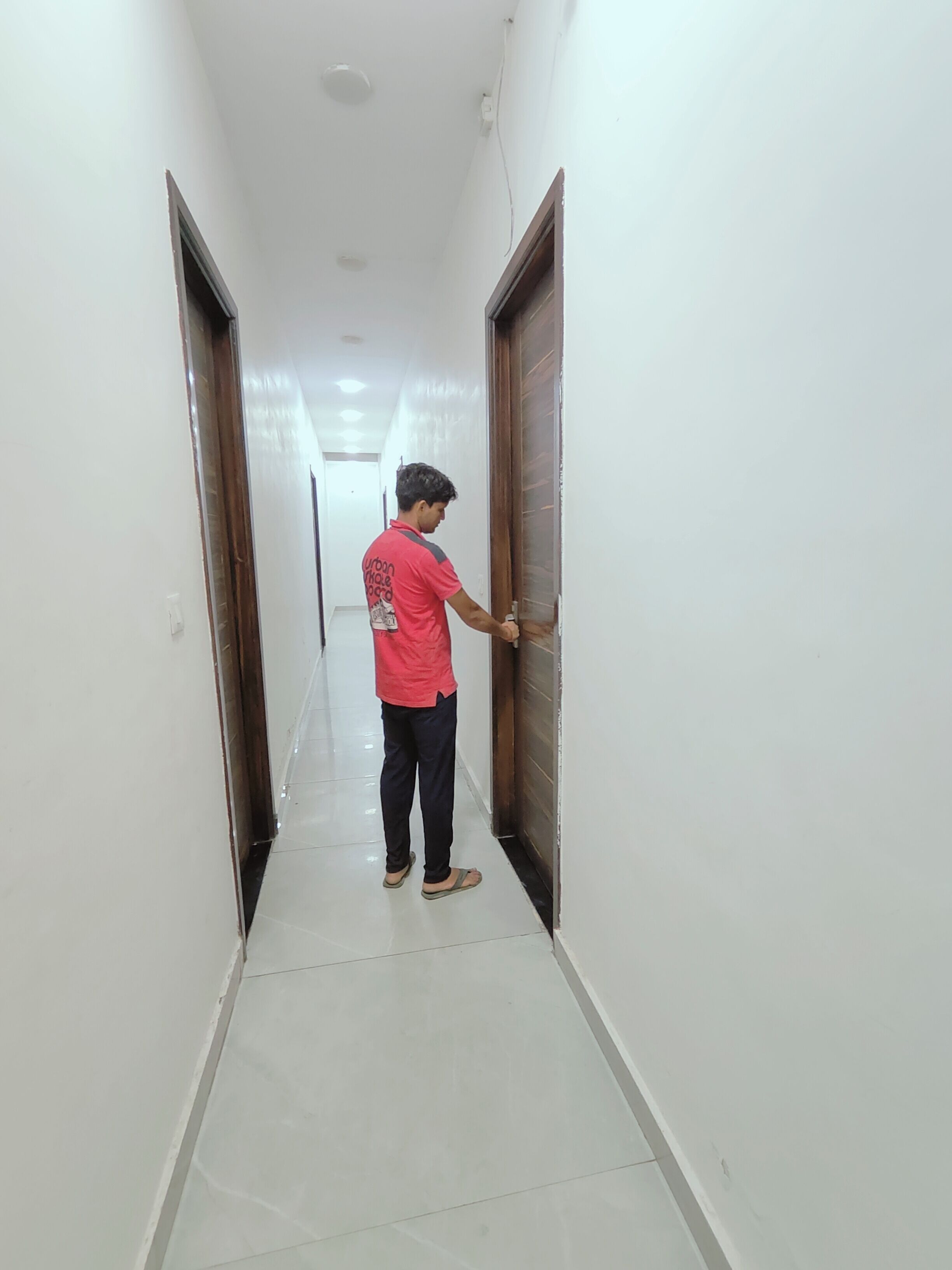 A person standing in a hallway with white walls and a tiled floor, doors on either side, and a ceiling light.