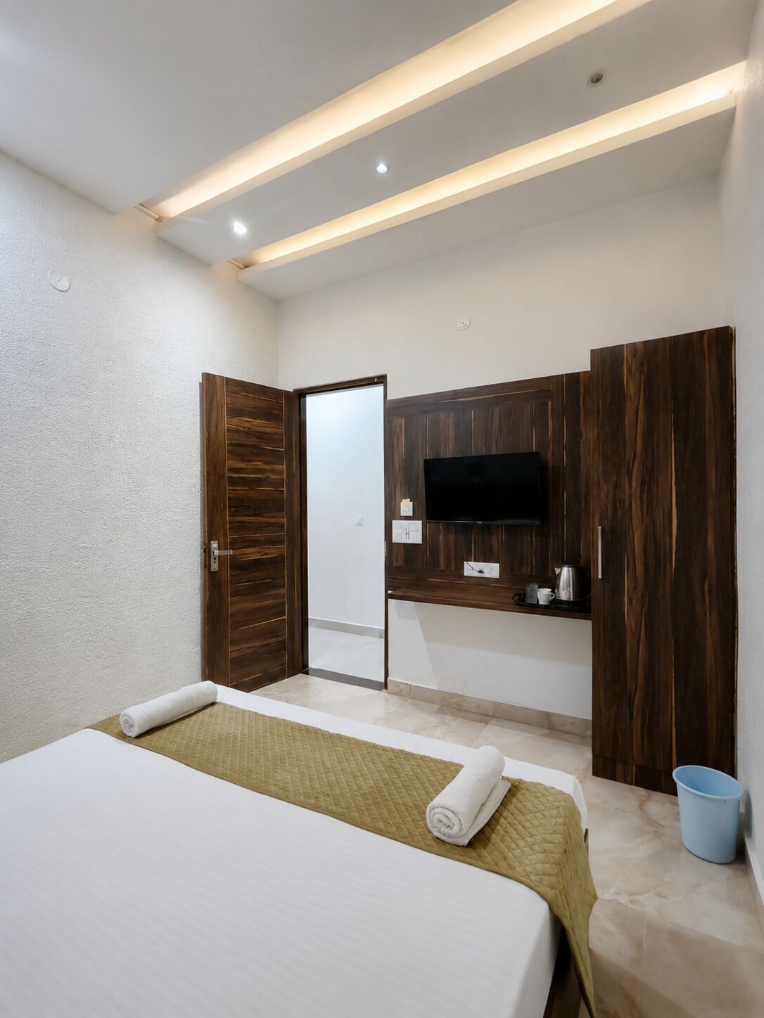 A hotel room with a bed, a TV mounted on a wooden panel, a small kitchenette, and a bathroom with a mirror.