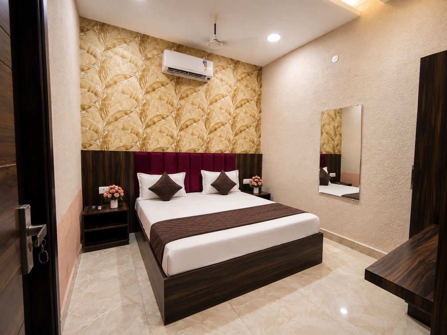 Habitación de hotel con una cama grande, aire acondicionado de pared, una mesita de noche con un jarrón de flores y un espejo en la pared.