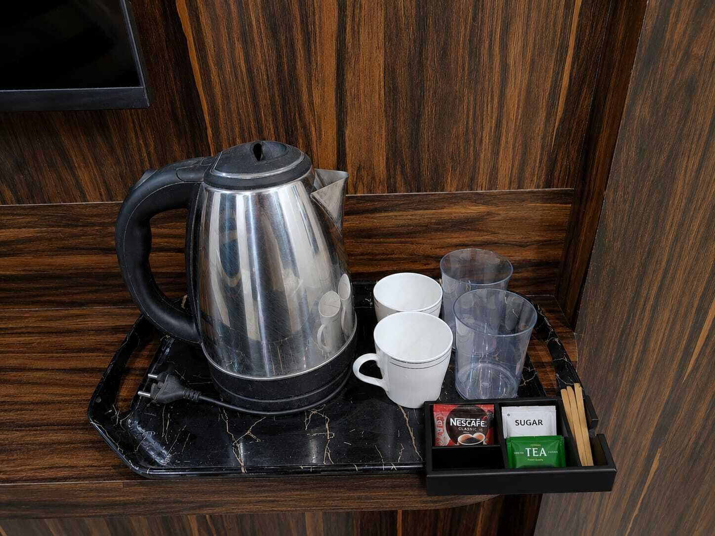 Una estación de café con una cafetera de acero inoxidable, tazas blancas, vasos de vidrio y una caja con Nescafé Classic, azúcar y té.