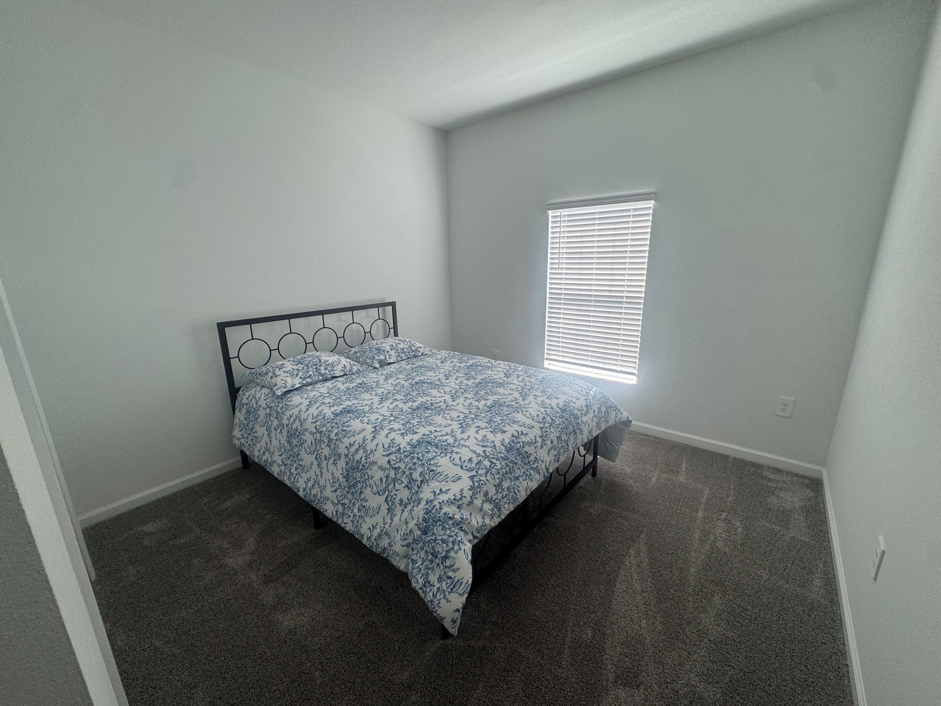 Una habitación pequeña con cama individual, cabecera sencilla, ventana con persianas y piso alfombrado.
