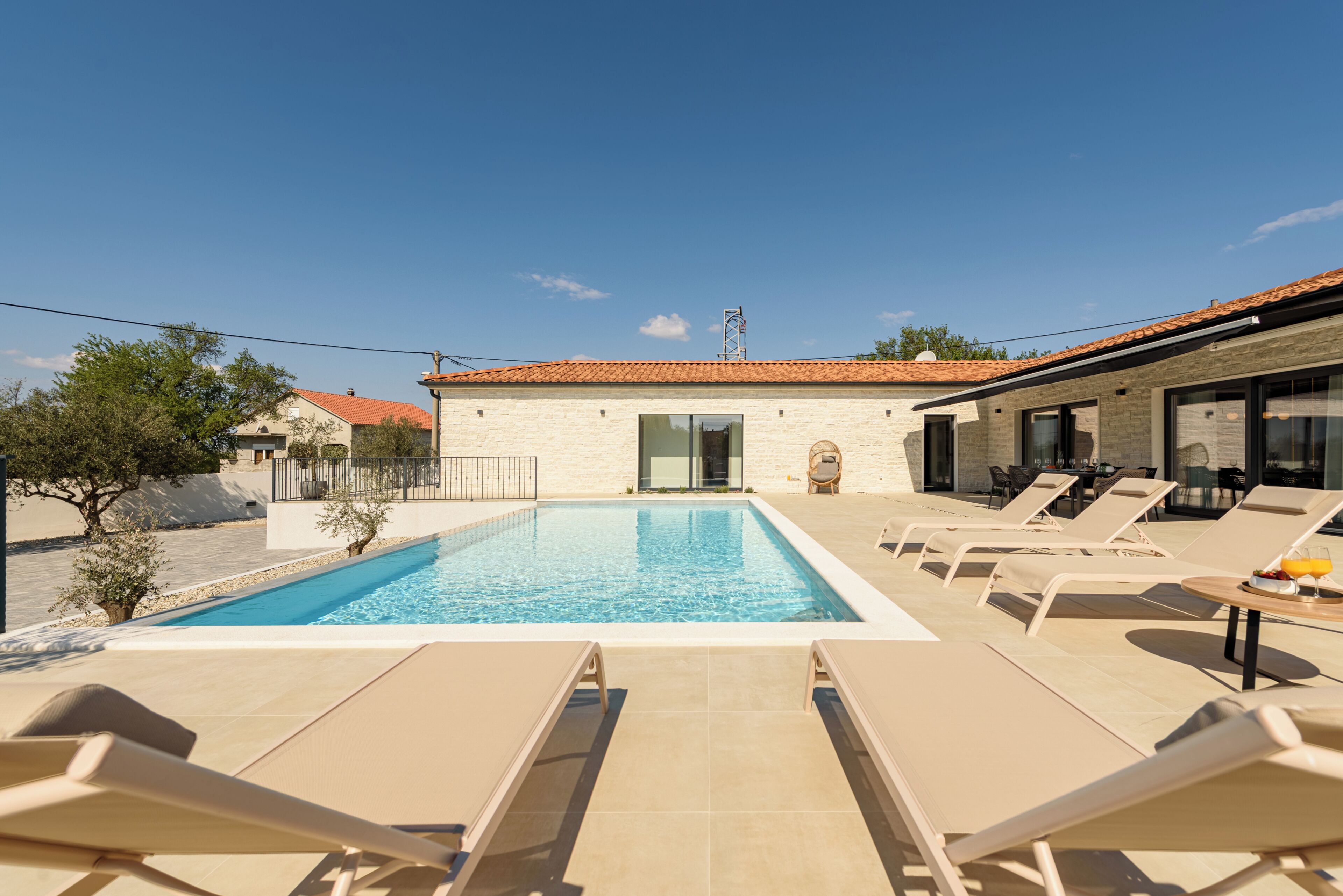 Une piscine avec des chaises longues, un bâtiment doté d’un toit en tuiles et un ciel dégagé.