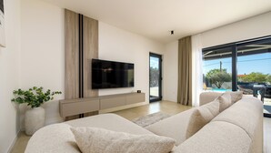 Una sala de estar moderna con un televisor de pantalla plana grande, un sofá color beige, una planta en maceta y vistas al exterior.
