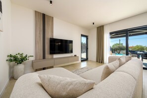 Ein modernes Wohnzimmer mit einem großen Flachbildfernseher, einem beigefarbenen Sofa, einer Zimmerpflanze und Blick ins Freie.