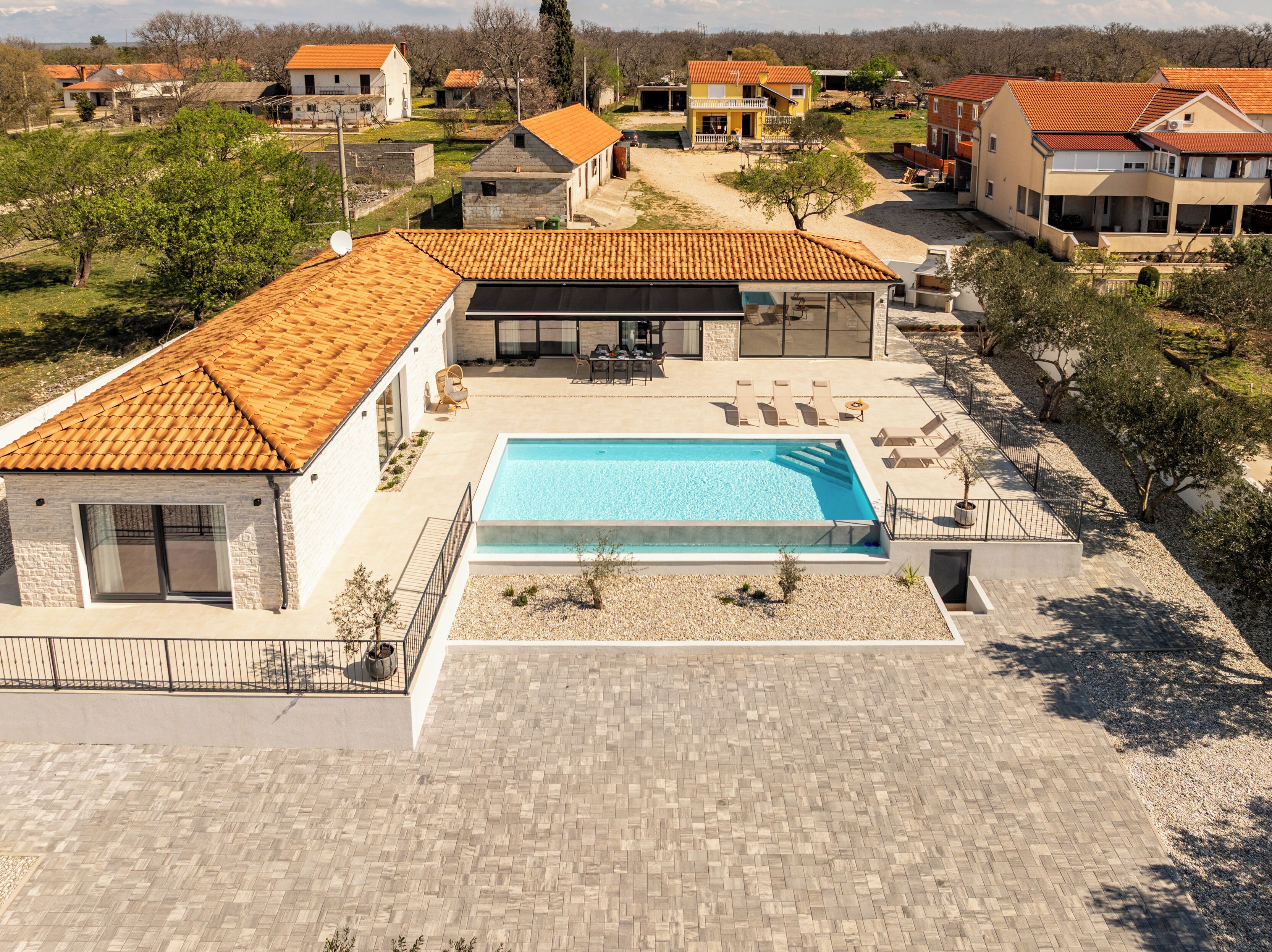 A residential area with a swimming pool, houses with orange tiled roofs, and a paved patio area.