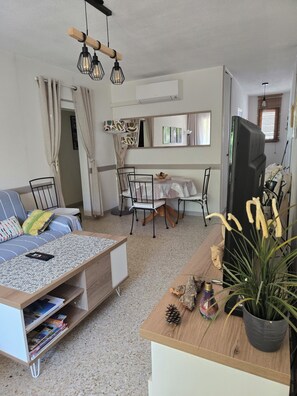 A living room with a sofa, dining table, chairs, a TV, and a potted plant. - Apartment T3 3*, 200m from the beach, 57m², 2 bedrooms, air conditioning, parking, terrace, balcony, shops (Bormes-les-Mimosas)