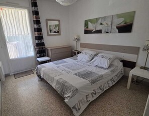 A bedroom with a bed, a bedside table, a nightstand, a lamp, a window with curtains, and a wall with a framed picture and a large flower painting. - Apartment T3 3*, 200m from the beach, 57m², 2 bedrooms, air conditioning, parking, terrace, balcony, shops (Bormes-les-Mimosas)