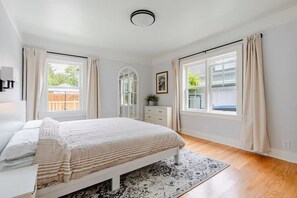 Ein Schlafzimmer mit Bett, Kommode, Spiegel und zwei mit Vorhängen versehenen Fenstern.