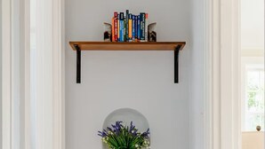 Un corridoio con una parete bianca, un pavimento in legno, una panca bianca, uno scaffale con libri e una pianta in vaso.
