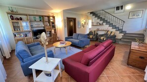 Ein Wohnzimmer mit einer Couch, einem Couchtisch und einem Fernseher. Eine Treppe mit Geländer und eine Tür zu einem anderen Raum sind ebenfalls vorhanden.
