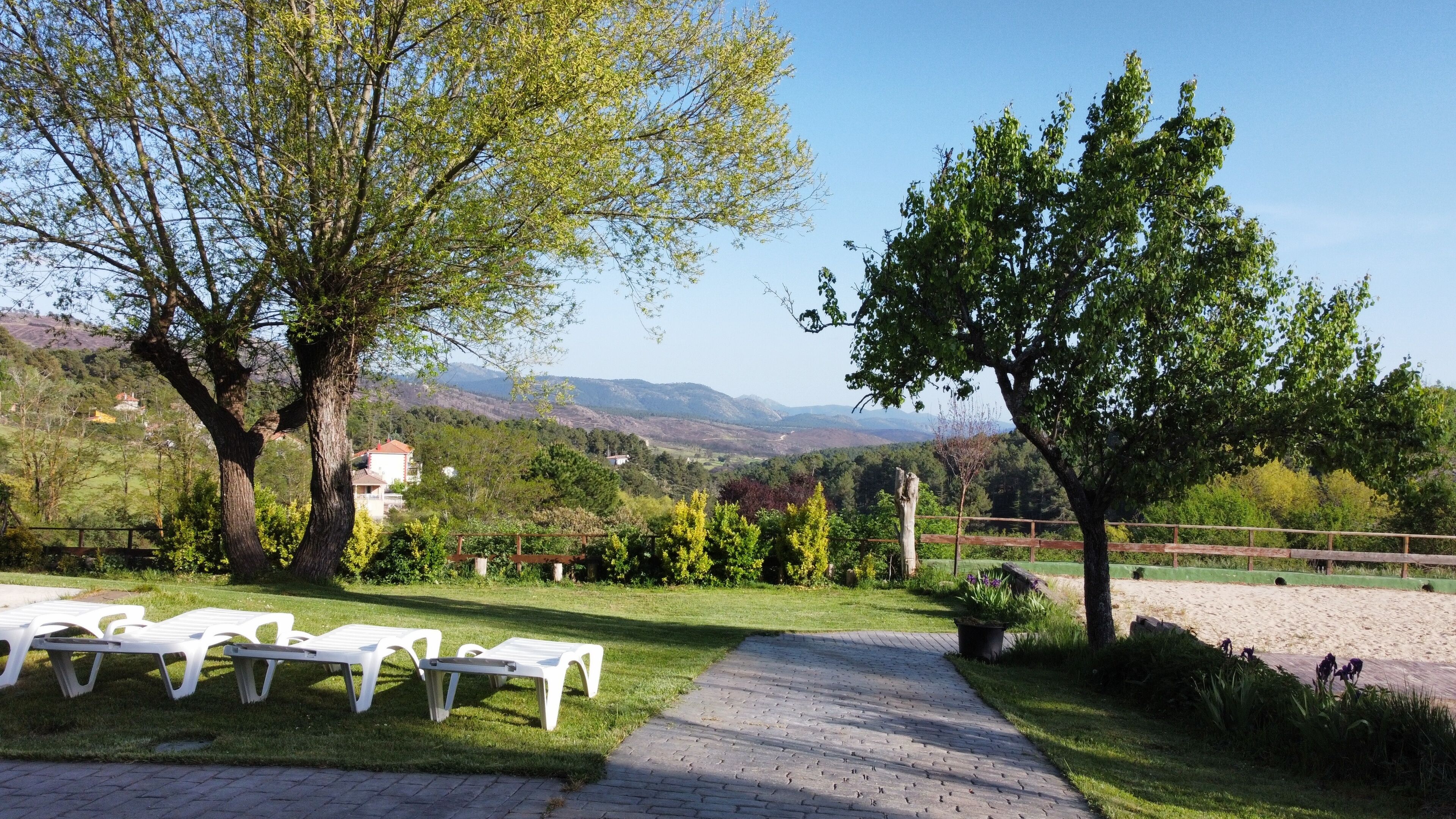 Un chemin pavé mène à des chaises longues blanches, entourées d’arbres et offrant une vue sur des collines lointaines.