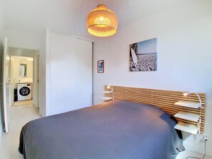 Ein Schlafzimmer mit einem Bett, einem Holz-Kopfteil, einer Wandlampe, einem Bild und einer Waschmaschine im angrenzenden Raum.