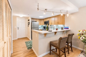 A compact kitchen with wooden cabinets, a stainless steel refrigerator, a microwave, and a bar area with stools. - Vega Apartment Seattle (Seattle)