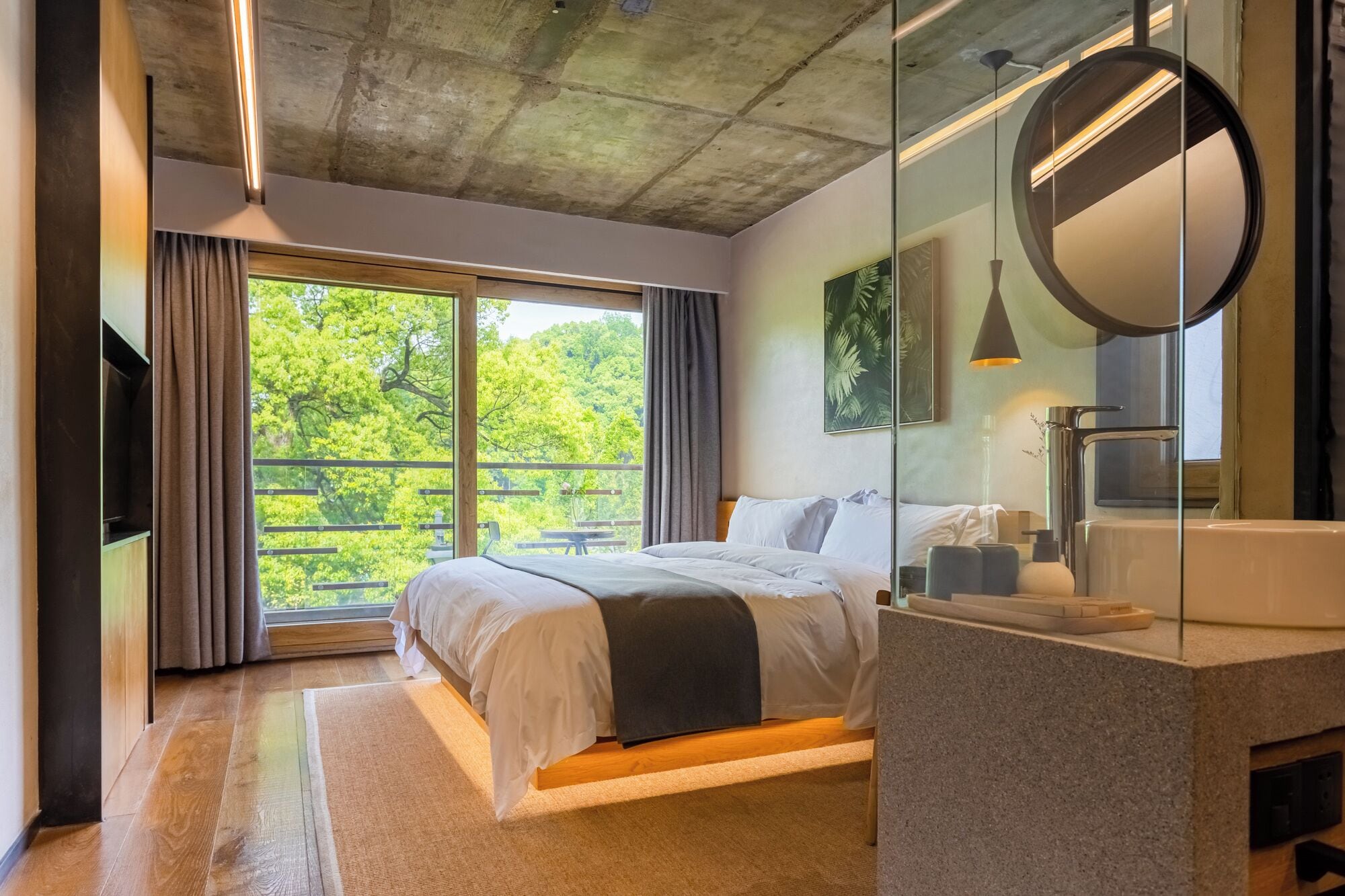 Une chambre moderne avec un grand lit, une cloison en verre, un miroir rond et une vue sur la verdure.