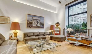 Ein modernes Wohnzimmer mit einer Couch, einem Glastisch mit Dekorationselementen, einer roten Lampe und einem großen Fenster mit Blick auf Gebäude.