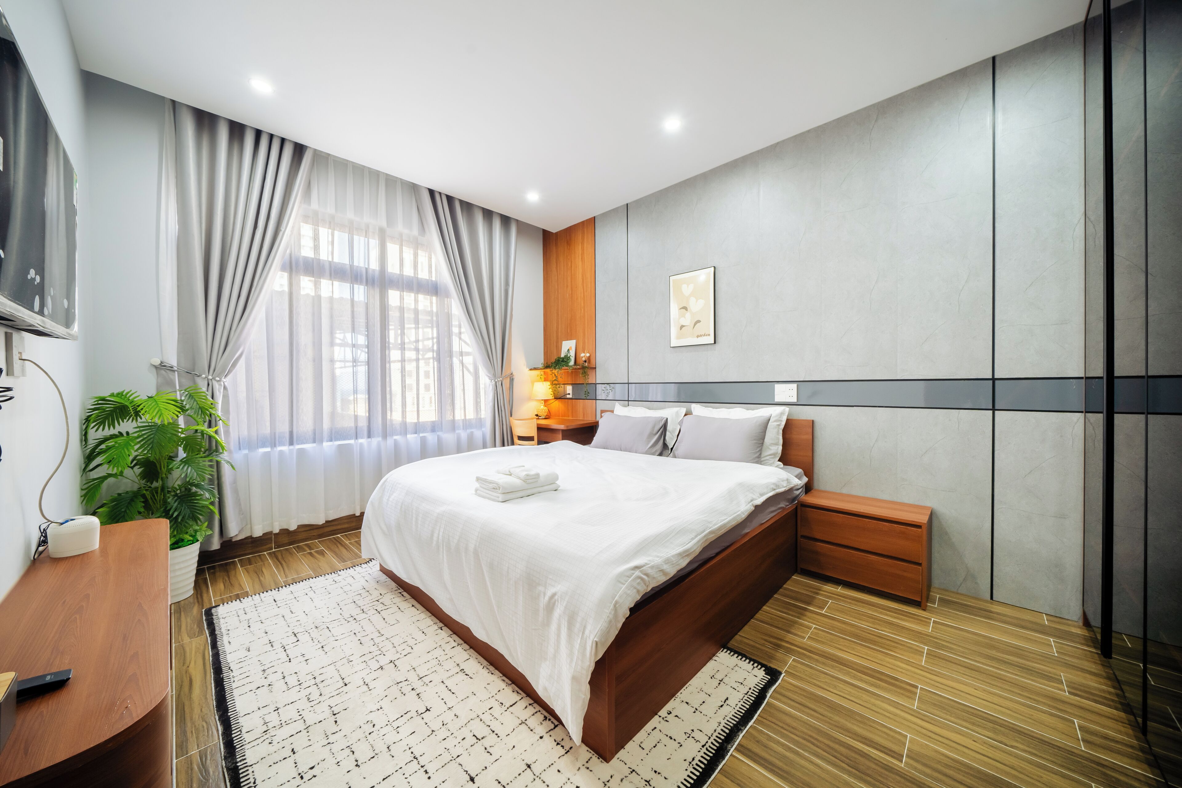 Ein modernes Hotelzimmer mit einem großen Bett, einem Nachttisch, einer Zimmerpflanze und einem großen Fenster mit Vorhängen.